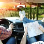 A man in a plaid shirt is driving an SUV into a sunset. A woman with her feet up on the dash is looking over a map.