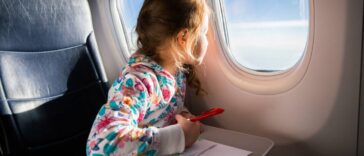 A young girl is sitting on an airplane and looking out the window. She is drawing a picture with colored pencils.