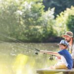 A father and son sitting next to each other on a pier and fishing. They're both holding a fishing pole in the water.