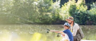 A father and son sitting next to each other on a pier and fishing. They're both holding a fishing pole in the water.