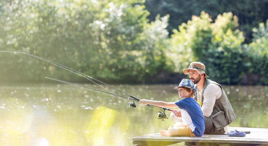 A father and son sitting next to each other on a pier and fishing. They're both holding a fishing pole in the water.