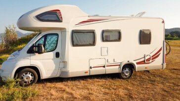 A white RV is parked outside in a field on a sunny day. The windshield and side windows are covered by sunshades.